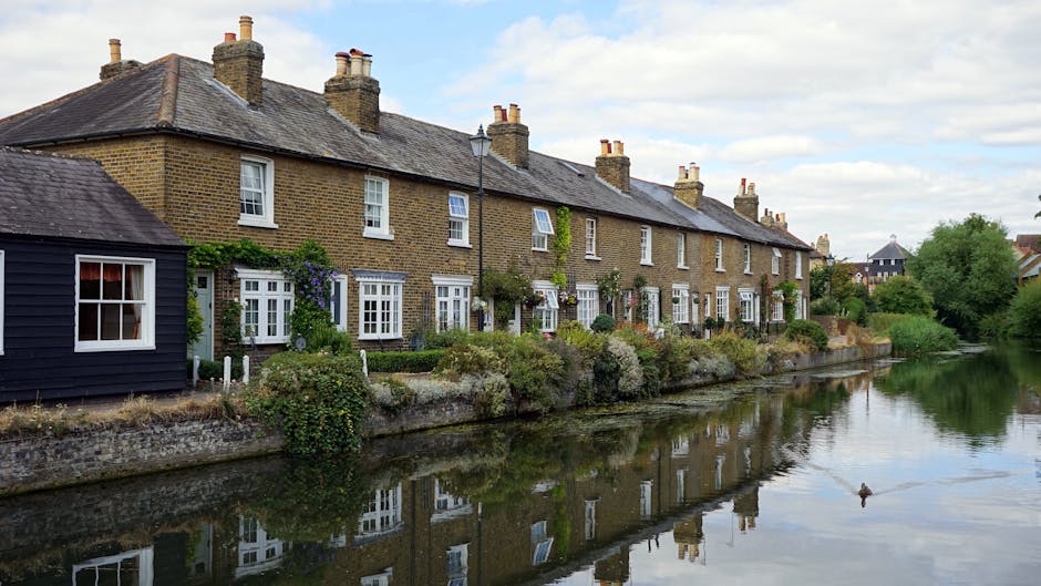 A row of traditional brick terraced houses with pitched roofs and multiple chimneys, situated along a canal in Cheam Village. The houses feature white-framed sash windows, some with small flower boxes