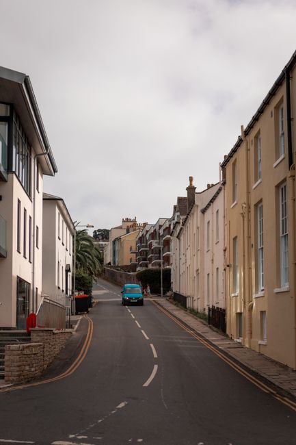 A narrow residential street with a gentle uphill slope, lined on both sides by closely spaced, multi-storey terraced houses painted in light, neutral colours such as cream and pale yellow. Several houses have large, vertically oriented windows, and some feature bay windows. The street surface is paved with asphalt and marked with a double yellow line along the edges, indicating parking restrictions. A single blue van is parked towards the middle of the street, likely being used for a home relocation or furniture transport as part of a house removal process. The area appears calm and quiet, with some greenery visible in the form of potted plants and small trees along the pavement, as well as a small palm tree near the steps on the left. The sky overhead is overcast, with diffused daylight providing natural lighting. This scene reflects an environment suitable for moving services such as packing, loading, and vehicle loading activities by companies like Man With a Van Sutton, operating in the vicinity of the St Helier estate, with emphasis on managing tight access areas for home removals.