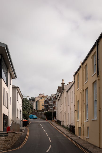 A daytime scene of a narrow residential street with a slight incline, featuring a row of attached cream-colored houses with white-framed windows and black window shutters on the right side, and modern white buildings on the left. The road is paved with asphalt, marked with double yellow lines along the edges, and has a dashed white line in the center indicating two-way traffic. In the distance, a man appears to be loading or unloading several cardboard boxes and wrapped furniture items from a dark-colored van parked near the curb, which is partially visible on the right. The street environment is lit by natural daylight, with an overcast sky overhead. The area is quiet, with no other vehicles or pedestrians present, suggesting a home relocation process involving furniture transport and packing, supported by [COMPANY_NAME], Man With a Van Sutton, specialising in removals and moving logistics in the area.