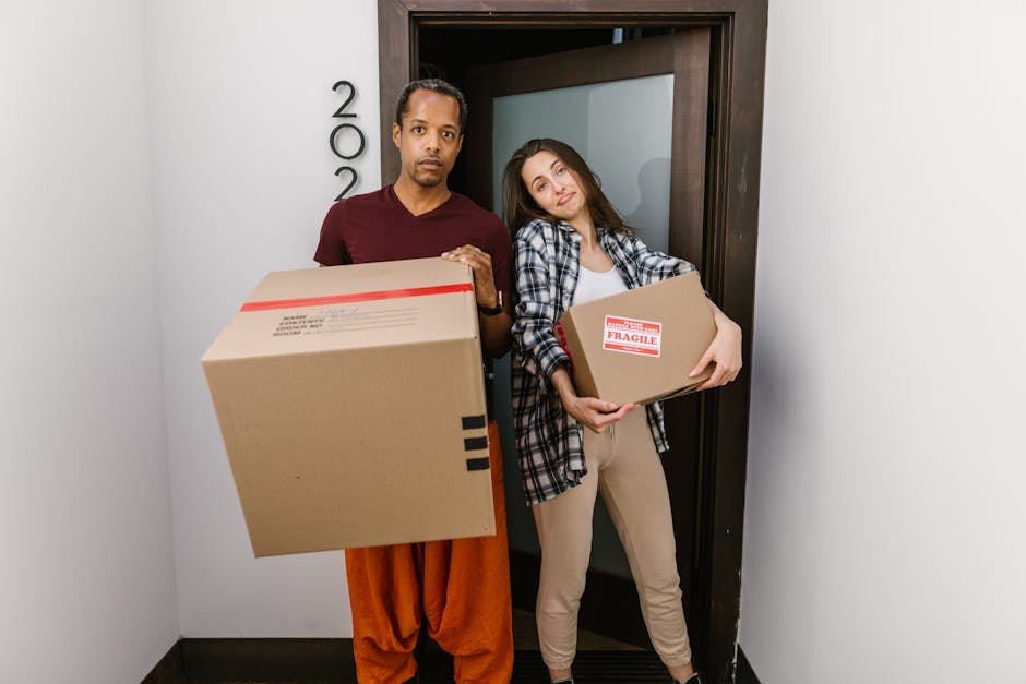 A man and a woman stand inside a doorway, each holding cardboard boxes associated with house removals. The man, with dark hair and wearing an orange pair of trousers and a dark red T-shirt, holds a large cardboard box in his hands, which is sealed with red tape and has a label indicating fragile contents. The woman, with long brown hair and wearing a plaid shirt over a white top and beige pants, carries a smaller cardboard box labeled 'FRAGILE' on the side. They are positioned in front of a dark wooden door with a metallic handle, and the room has plain white walls. The natural lighting enhances the clarity of packaging materials such as cardboard, tape, and the boxes’ labels, suggesting a home relocation scenario involving packing, loading, and furniture transport. This image represents the work of a professional removals service like Man With a Van Sutton, highlighting the typical steps of packing and moving within a house or apartment, including careful handling of fragile items during the loading process.