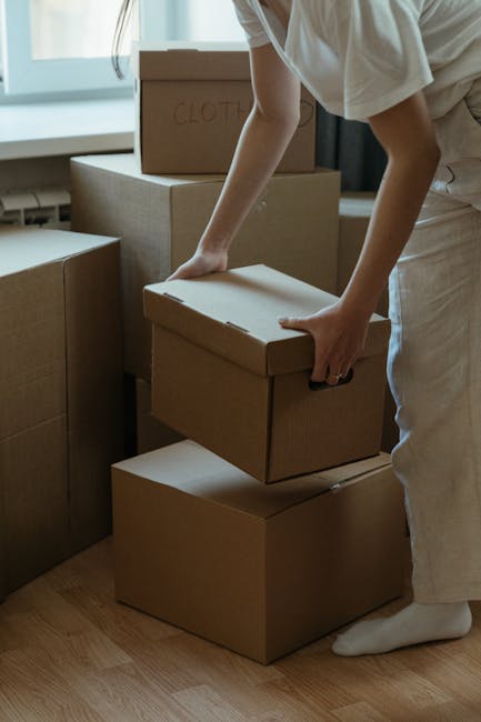 A person, partially visible and dressed in light-colored clothing, is seen stacking cardboard boxes in an indoor setting, likely a home or storage area, with a window allowing natural daylight to illuminate the space. The boxes vary in size, with some labeled and wrapped in packing materials; the individual is carefully placing a box on top of other boxes arranged near the window. In the background, larger boxes are stacked, and some are reinforced with tape, indicating packing for a home relocation or furniture transport process. The floor is wooden, and the scene suggests an organized packing and moving sequence, typical of house removals. As part of professional moving services provided by Man With a Van Sutton, this loading process illustrates careful handling of household items, including delicate or boxed possessions, in preparation for transportation to a new residence or storage facility, aligning with services such as furniture removals and packing during house moves.