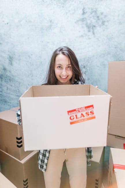 A man and a woman stand inside a doorway, each holding cardboard boxes associated with house removals. The man, with dark hair and wearing an orange pair of trousers and a dark red T-shirt, holds a large cardboard box in his hands, which is sealed with red tape and has a label indicating fragile contents. The woman, with long brown hair and wearing a plaid shirt over a white top and beige pants, carries a smaller cardboard box labeled 'FRAGILE' on the side. They are positioned in front of a dark wooden door with a metallic handle, and the room has plain white walls. The natural lighting enhances the clarity of packaging materials such as cardboard, tape, and the boxes’ labels, suggesting a home relocation scenario involving packing, loading, and furniture transport. This image represents the work of a professional removals service like Man With a Van Sutton, highlighting the typical steps of packing and moving within a house or apartment, including careful handling of fragile items during the loading process.